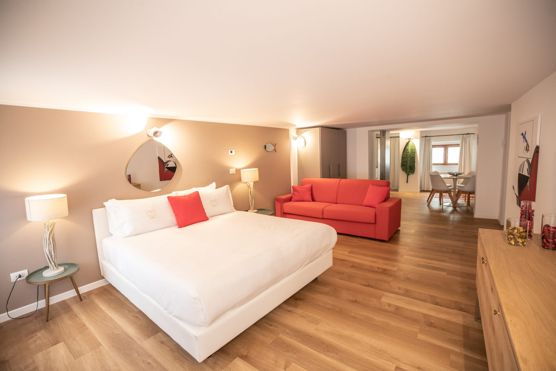 Stanza spaziosa con un letto matrimoniale, un divano rosso, specchio rotondo, lampade da terra e una vista sulla sala da pranzo.