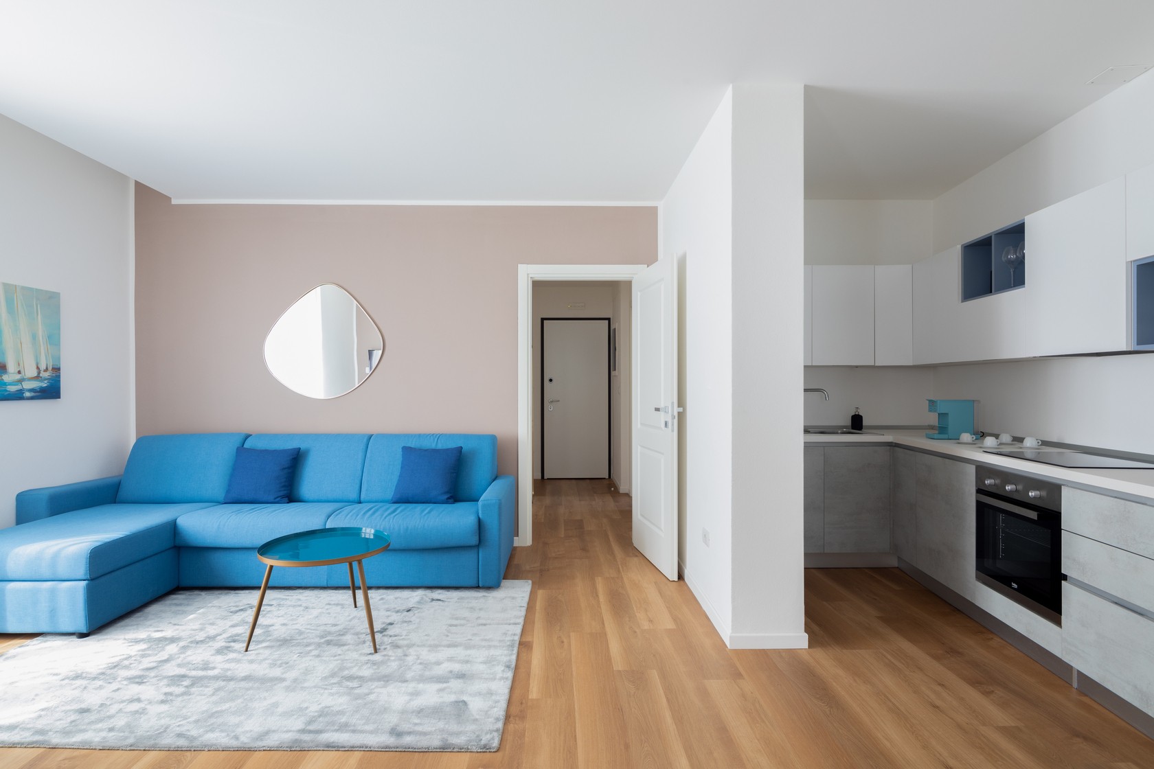 L'immagine mostra un soggiorno con cucina a vista. C'è un divano blu, un tappeto grigio, un tavolino e uno specchio a forma di goccia.