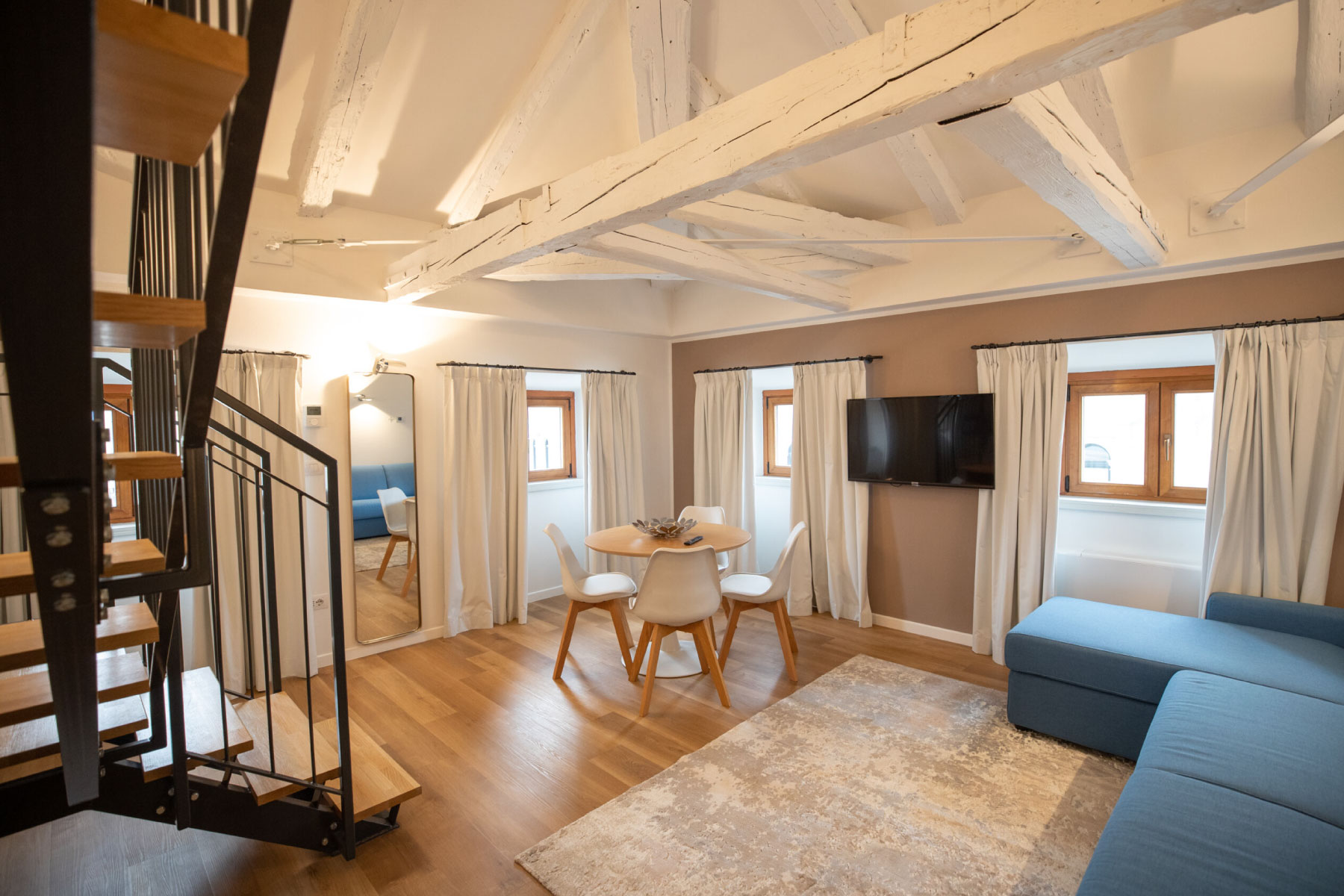 Ampio soggiorno con divano blu, travi in legno bianche a vista, tavolo da pranzo rotondo, sedie bianche e scala a chiocciola nera.